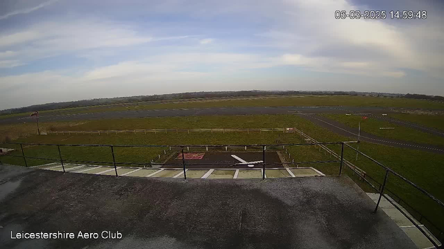 A wide view of an airport runway on a clear day. In the foreground, there is a flat rooftop with a railing. The runway stretches into the distance, surrounded by green grass and a few light-colored barriers. A small building is visible on the right side of the image, with a parking area and multiple taxiways marked on the pavement. The sky is mostly clear with a few thin clouds, and a small flag is visible on a pole near the left side of the frame. The date and time are displayed in the top right corner.