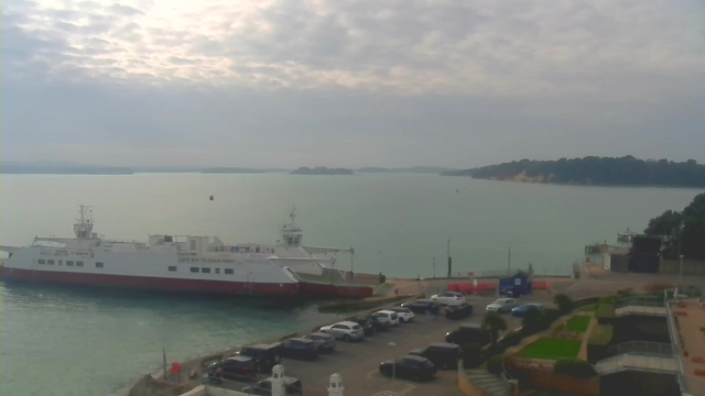 A ferry boat is docked at a pier, with a calm blue-green sea and a cloudy sky. Several parked cars are visible in a parking lot next to the pier, and there are some plants and steps leading to a lower area. In the background, a shoreline with greenery and distant hills can be seen.