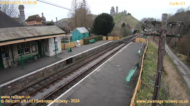 A view of a railway station at Corfe, featuring a stone platform with green benches and a small building with a slate roof. In the background, there are wooden fences and a green shed. The railway tracks run parallel to the platform, leading off to the right. A historic castle ruins can be seen atop a hill in the distance, surrounded by trees and grassy slopes. The sky is overcast.