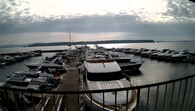 A view of a marina filled with various boats docked at a wooden pier. The scene is set under a cloudy sky, with soft sunlight breaking through the clouds, reflecting on the water. In the foreground, a large white yacht is prominent, surrounded by smaller boats of different shapes and sizes. The background includes a distant shoreline and more boats arranged neatly along the water.