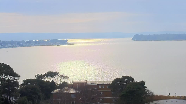 A serene view of a large body of water, reflecting soft sunlight. In the foreground, there are silhouettes of trees and a low building, blending into the landscape. The background features a distant shoreline with some buildings and gentle hills, under a pale cloudy sky. The overall atmosphere is calm and peaceful, evoking a sense of tranquility by the water.