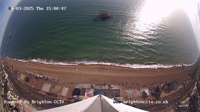 Aerial view of a beach with golden sand and rolling waves meeting the shore. There are various colorful beach structures and attractions visible near the water, including people walking along the beach. In the distance, a pier extends into the ocean, and a structure, possibly a derelict pier, is visible partially submerged in the water. The sky is bright with sunlight reflecting off the water surface, indicating a clear day. The date and time are displayed at the top of the image.