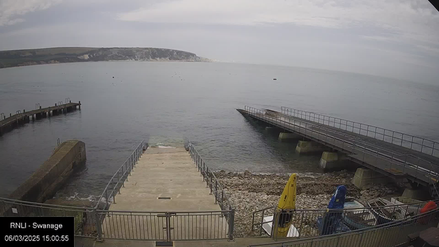 A view of a calm sea with patches of land in the background. In the foreground, there is a concrete ramp leading down to the water, bordered by a metal railing. To the left, a stone wall juts out into the water. Along the right side, there are two boat slips with boats parked nearby, including a yellow kayak. The sky above is mostly overcast with soft clouds. The scene appears tranquil, with gentle waves lapping at the shore.