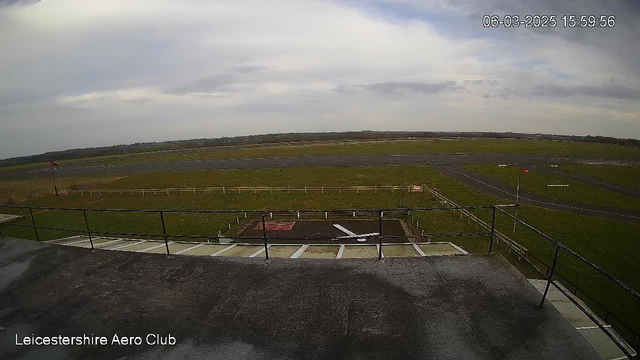 A view from a webcam at Leicestershire Aero Club showing a grassy airfield under a cloudy sky. In the foreground, there is a flat rooftop with a railing. Below, the airfield is marked by a large expanse of green grass intersected by runways made of dark asphalt. A windsock stands to the left, and there are multiple small markers on the runway. The scene is tranquil with no aircraft visible at this time. The timestamp indicates the image was captured on March 6, 2025, at 15:59:56.