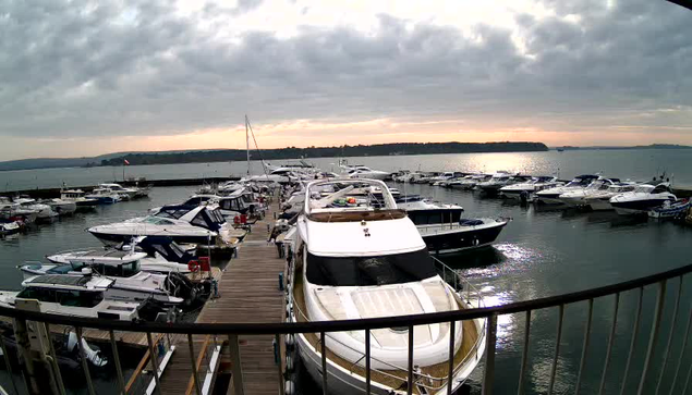 A view of a marina with several boats docked at wooden slips. In the foreground, a large white motor yacht is prominent, with a canopy on top and various items on the deck. The water is calm, reflecting the cloudy sky above. A distant shoreline is visible in the background, with trees and hills. The overall scene is tranquil, with muted colors and a hint of sunlight breaking through the clouds at the horizon.