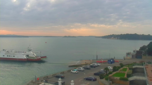 A view of a calm harbor at dusk. A ferry with a red and white design is navigating the water, while several parked cars are visible along the shoreline. In the background, rolling hills and trees can be seen, silhouetted against a cloudy sky. The water reflects soft hues of orange and gray from the fading sunset. To the right, there's a small building and some landscaped areas.