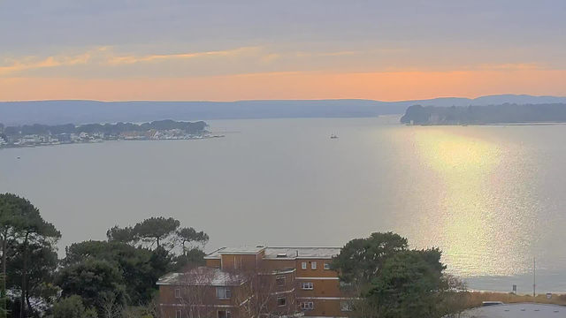 A serene waterfront scene is depicted with calm water reflecting soft hues of orange and pink from the sunrise. In the foreground, there are trees and a brown building partially visible. Further out, small boats can be seen on the water, leading to the horizon where a range of blue hills meets the sky. The overall atmosphere is tranquil and picturesque, suggesting a peaceful morning by the water.