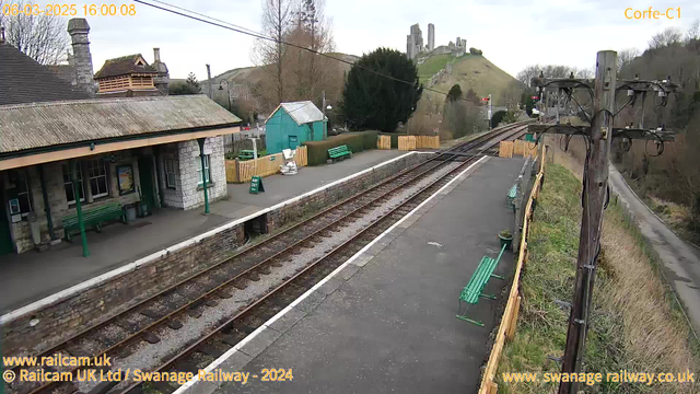 A view of a railway station with a partially covered platform featuring several green benches. On the left, there is a stone building with a sloped roof, and behind it, a small turquoise shed. The railway tracks run parallel to the platform, leading towards a hillside in the background where a castle ruins can be seen atop. Several trees and shrubs are scattered around, and a wooden fence borders the area. A power pole with wires stands on the right side of the image. The sky is cloudy, and the timestamp shows the date and time.