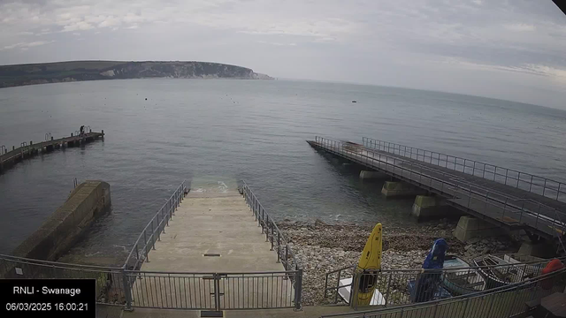 A view of a calm sea with a cloudy sky overhead. In the foreground, there are two loading ramps leading to the water. On the left, a stone jetty extends into the sea, while on the right, a wooden pier is visible. Several boats are moored in the water, and there are kayaks in bright colors, including yellow and blue, arranged on a stony shoreline. The scene is serene, evoking a quiet coastal setting.