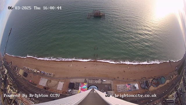 Aerial view of a coastal area, showing a sandy beach with waves lapping at the shore. There are a few people walking on the beach and some distant figures in the water. A partially submerged pier is visible in the ocean. The beach includes colorful beach huts, a water pool, and a Ferris wheel. The timestamp in the corner indicates the date and time as 06-03-2025, 16:00:44. The bottom of the image has text indicating it is powered by Brighton CCTV.