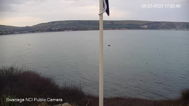 A coastal view captured from a webcam, showing calm waters with a slight ripple. In the foreground, there is sparse vegetation and some rocky terrain. The background features a town along the shoreline, with buildings visible. Gentle hills rise behind the town, leading to a cloudy sky overhead. In the top right corner, a timestamp reads "06-03-2025 17:00:46." A flagpole stands on the left side of the image.