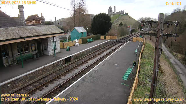 A webcam image of a railway station platform at Corfe Castle. The scene shows a long, rectangular platform with a textured gray surface, bordered by wooden fencing on the right. Green benches, some facing the tracks, are scattered along the platform. In the background, a small green shed is visible, and a "WAY OUT" sign is placed near the platform edge. The hillside behind the station features ruins of a castle, partially obscured by trees. The sky is overcast, and the time displayed in the corner is 16:59:50 on March 6, 2025.