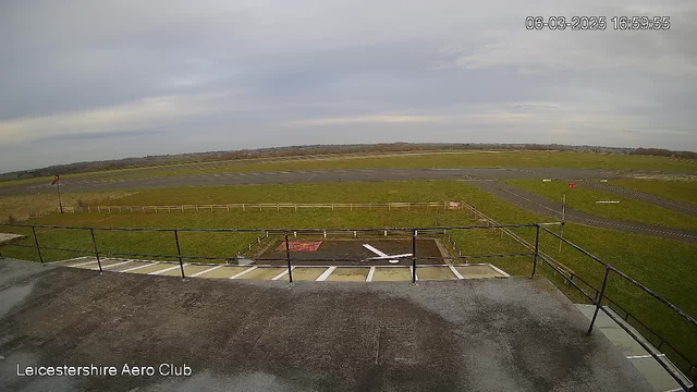 A wide view of an aerodrome, taken from a raised platform. The foreground features a railing, with a concrete surface beneath. In the background, a large grassy area leads to a tarmacked runway. The runway is bordered by a wooden fence. A small windsock is visible to the left, and there are markings on the runway. The sky is overcast with a light gray hue, and the overall lighting suggests a daytime setting. An indication of the date and time is visible in the top right corner.