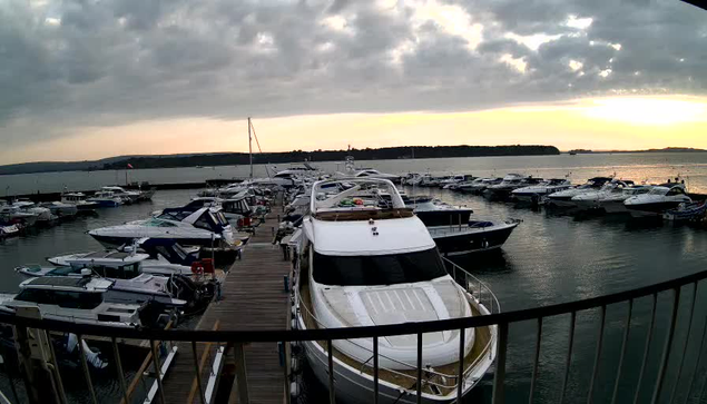 A view of a marina at sunset, with numerous boats docked. In the foreground, there is a large white yacht, prominently positioned among smaller vessels. The water reflects the soft colors of the sky, which features a mix of clouds and open space, indicating evening light. A wooden dock leads towards the boats, with a railing visible in the lower part of the image. The horizon is lined with land in the distance, adding depth to the scene.