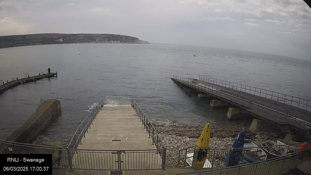 A view of a calm sea under a cloudy sky, with a rocky shoreline and steps leading down to the water. In the distance, cliffs can be seen on the opposite side of the bay. A wooden pier extends into the water, and there are some kayaks, including a yellow one, resting near the shore. The image has a timestamp and location label at the bottom.