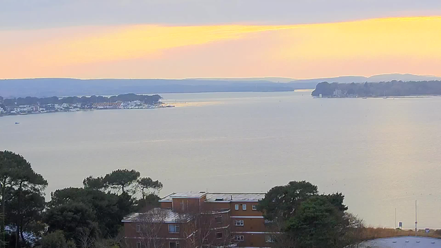 A panoramic view of a calm body of water at sunset. The sky features soft hues of orange and pink, transitioning into blues. On the left, a small marina with boats is visible next to a line of buildings. In the foreground, there are trees framing the view, while the water stretches out toward distant hills and land masses on the right. The scene conveys a tranquil and serene atmosphere.