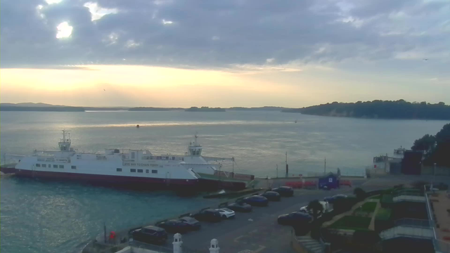 A ferry boat is docked at a pier with a backdrop of calm water and a cloudy sky. The horizon features faint hills silhouetted against the setting sun, which casts a warm glow over the scene. In the foreground, several parked cars are visible along the waterfront, and there are patches of greenery and landscaped areas beside the pier. The overall atmosphere is tranquil, with gentle water ripples reflecting the cloudy sky.