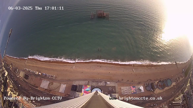 Aerial view of a sandy beach with waves lapping at the shore, featuring a pier extending into the water on the left side. A dilapidated structure is partially submerged in the sea. The beach is lined with an amusement park, colorful structures, and a promenade. The sun casts a bright glare on the water in the upper right. The image includes a timestamp indicating the date and time.