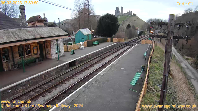 An image of a train station platform at dusk. The foreground shows a concrete platform with several green benches along the edge. On the left, there is a stone building with a sloped roof, featuring large windows and a sign partially visible that reads “Corfe Castle.” A blue shed is situated slightly behind the building. The platform extends to the right, where train tracks run parallel, leading towards a distant hillside. In the background, ruins of Corfe Castle are visible atop the hill. A wooden utility pole with wires is positioned at the far right of the image.