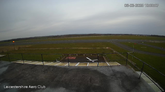 A view from a rooftop at Leicestershire Aero Club, overlooking an airfield. The foreground features a balcony railing and a small helicopter landing area with a white airplane silhouette. In the background, the airfield consists of grass and asphalt runways, with a few marked areas and a flag in the distance. The sky is overcast, creating a gray atmosphere, and there is a dimly lit orange beacon on the left. The time is displayed as 18:00 on June 3, 2025.