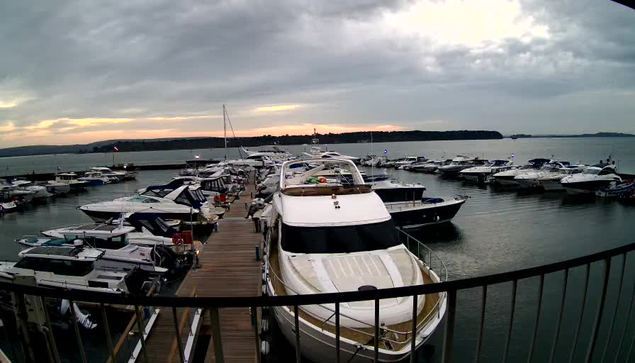 A marina scene with numerous boats docked in calm water. The boats vary in size and style, with some featuring bright blue and white colors. A wooden walkway runs along the dock, leading to a sunset in the background that casts a soft light across the sky, which is filled with clouds. The water reflects the muted colors of the overcast sky and the silhouette of distant land can be seen on the horizon.