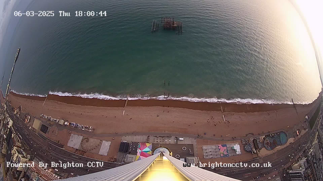 A view from a high vantage point looking down at a sandy beach along a coastline. The beach has a few scattered beachgoers and sections of blue water with white waves gently lapping at the shore. There is a dark structure partially submerged in the water a short distance from the shore. On land, there are amusement rides and colorful structures, with a bright pattern visible on one of the buildings. A long pier extends into the water, and the scene captures the golden hue of the setting sun, suggesting late afternoon. The image includes a timestamp and a watermark at the bottom left.