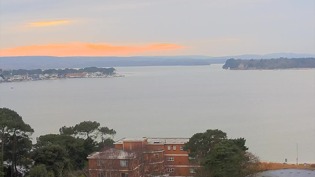 A tranquil waterfront scene is depicted, with a calm body of water reflecting the pale sky. In the background, there is a distant shoreline with trees and buildings lining the water's edge. The foreground features a brick building partially obscured by greenery. A soft orange glow on the horizon indicates a sunrise or sunset, while gentle hills are visible in the distance. The overall atmosphere is serene and natural.