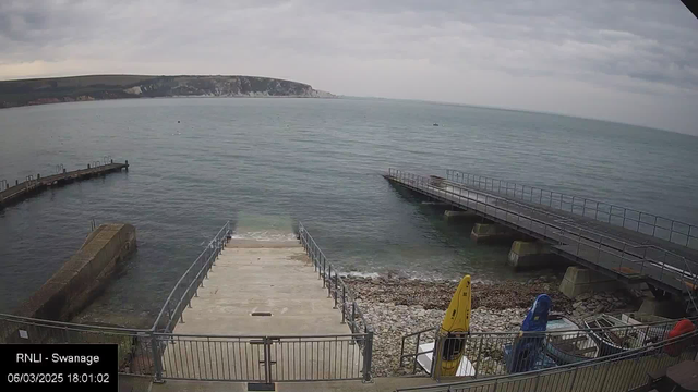 A view of a calm sea under a cloudy sky. In the foreground, there is a concrete ramp leading down to the water, bordered by a metal railing. To the right, there are two jetties extending into the water, one with a wooden surface. On the shore, there are some boats, including a yellow kayak and a blue kayak, along with some rocky beach area. In the background, a cliff can be seen along the shoreline.