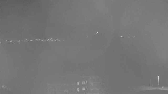 A night view captured from a webcam, primarily in shades of gray. In the foreground, a dimly lit building can be seen, with a few windows appearing slightly illuminated. The background features a skyline with scattered lights, indicating distant structures or roads. The overall ambiance is dark, with limited visibility due to a hazy atmosphere.