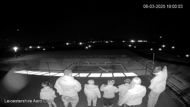 A group of five people stands together on a balcony at night, overlooking an area with lights in the distance. The individuals include two adults and three children. One adult points towards something in the distance. The balcony has a railing, and in front of the group, a light-colored structure is visible. The scene is mostly dark, with the main source of illumination coming from the distant lights. The date and time are displayed in the upper right corner.