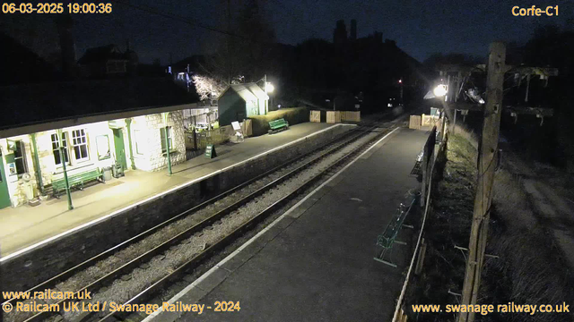 A dimly lit railway station at night. The platform features a stone building with green railings and benches. A sign reads "WAY OUT" near a wooden fence. Train tracks run alongside the platform, with a few benches on either side. Surrounding trees and buildings are silhouetted against the night sky.