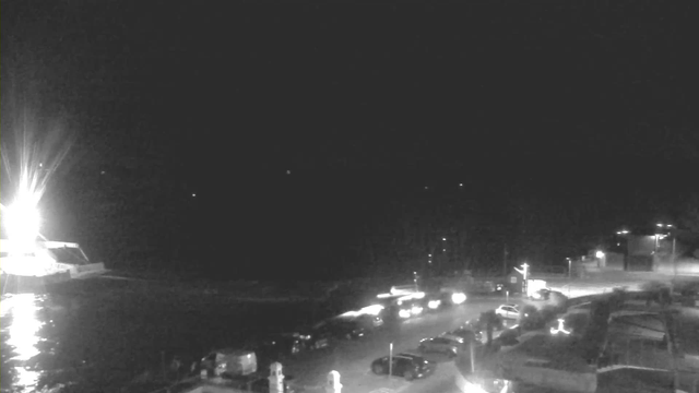 A nighttime view of a road along the coast, illuminated by various lights. A boat is visible in the water to the left, with a bright light source shining from it. On the road, several parked vehicles can be seen along with some moving cars. The background features a dark sky, hinting at a calm night with a few distant lights visible over the water. The scene conveys a tranquil coastal atmosphere.