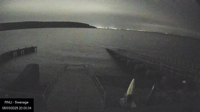 A dimly lit scene of a harbor area at night. In the foreground, a wooden pier juts out into a calm body of water. To the right, several small boats are visible, one with a white sail. The background features a distant shoreline illuminated by lights, likely from a town or city. The sky is overcast with clouds, adding to the moody atmosphere of the scene.