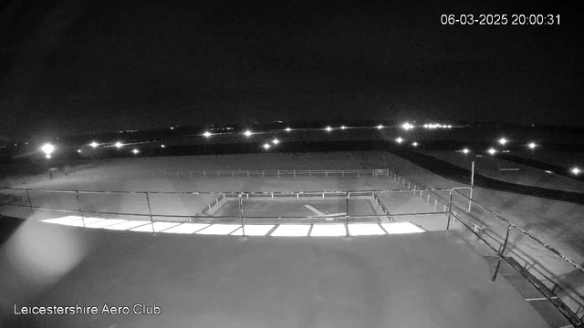 A black and white image capturing a nighttime scene at the Leicestershire Aero Club. The foreground shows a fenced area with some visible ground and light areas reflecting under a railing. In the background, several lights are scattered across the darker landscape, likely indicating nearby structures or roadways. The date and time are displayed in the upper right corner, showing March 6, 2025, at 8:00 PM.