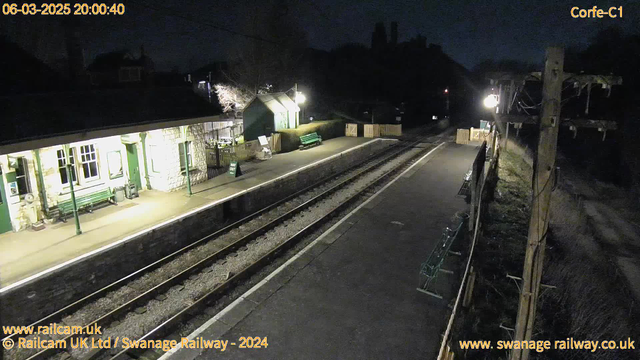 A nighttime scene at a railway station. The platform is illuminated by bright lights, showing multiple green benches on a stone pathway beside two parallel railway tracks. On the left, there is a small building with a porch, featuring large windows and a green gate marked "Way Out." To the right, there is a wooden fence and more benches, with a distant view of trees silhouetted against the dark sky. The atmosphere is quiet and still, typical of a nighttime railway station.