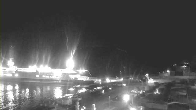 A nighttime scene at a waterfront location. In the foreground, there are several boats docked along the shore, illuminated by nearby lights. A large ferry-like boat is anchored on the left side of the image with bright lights on its deck creating a glowing effect. The water reflects the lights, creating a shimmering surface. The background shows a few more lights from structures along the shore, indicating a busy but tranquil harbor area.