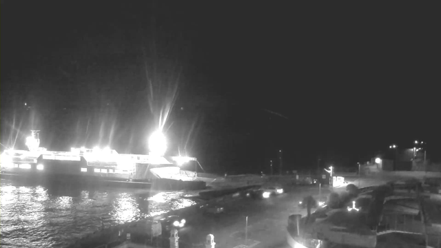 A nighttime scene of a harbor with a large boat docked. The boat is brightly illuminated, reflecting light on the water. In the foreground, there are several cars on a road next to the water. The setting is relatively dark, with additional faint lights in the background, possibly from other buildings or distant vehicles. The overall atmosphere is calm and quiet, typical of a port area at night.