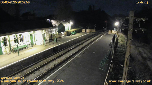 A dimly lit train station at night. The platform has wooden benches and is lined with low stone walls. On one side, there is a small building with large windows and green decor. A green sign indicates an exit path, and a lamppost provides soft lighting. In the background, shadows of trees and buildings are present, adding to the nighttime atmosphere. Two railway tracks run along the platform, and the scene is mostly empty and quiet.