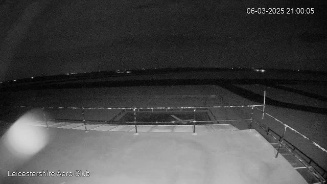 A dark nighttime scene from a webcam at the Leicestershire Aero Club. The image shows a flat rooftop with a railing visible at the front. In the background, faint lights from the horizon indicate a distant landscape. The ground appears flat and expansive, while the sky is mostly clear, indicating nighttime. The timestamp in the corner reads March 6, 2025, at 21:00:05.