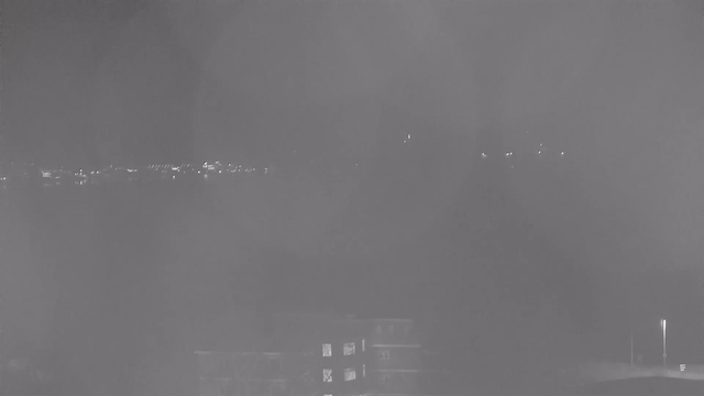 A low-light webcam image showing a hazy view of a cityscape at night. In the distance, there are faint points of light that appear to be streetlights or building lights. The foreground features a dark outline of structures, one with illuminated windows, while the background is mostly dark, hinting at a skyline. The overall image has a monochromatic tint, with varying shades of gray.