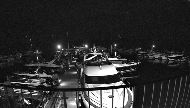 The image shows a marina at night, filled with various boats and yachts docked along wooden piers. Some boats have lights illuminating them, while others are in shadow. The scene is predominantly dark, with a few bright spots where nighttime lights are reflecting on the water. The perspective suggests a view from a higher platform.