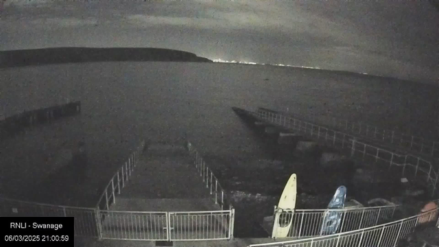 A dimly lit waterfront scene at night. A pier extends into the dark water, with several boats moored alongside. There are two canoes on the shore, one yellow and one blue, leaning against a white railing. The water appears calm, while faint lights from a distant shoreline are visible across the water. The sky is mostly cloudy, contributing to the low light conditions.