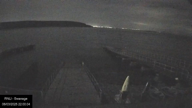 A dark, nighttime scene of a calm body of water with distant lights visible on the horizon. In the foreground, there are wooden piers extending into the water. Slightly to the right, a small sailboat is partially visible, sitting on the dock. The overall atmosphere is quiet and serene, with a cloudy sky above.