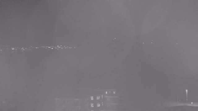 A dimly lit scene captured by a webcam at night. The foreground contains dark shapes that suggest buildings, while blurry lights from the background indicate a distant cityscape or shoreline. The overall image has a smoky or hazy appearance, making details hard to discern.