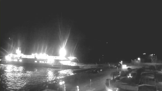 A dimly lit scene showing a large ferry docked at a port. Bright lights illuminate the ferry, reflecting off the dark water. The dock area is visible with parked vehicles and faint outlines of structures, while the background remains mostly dark, suggesting nighttime.