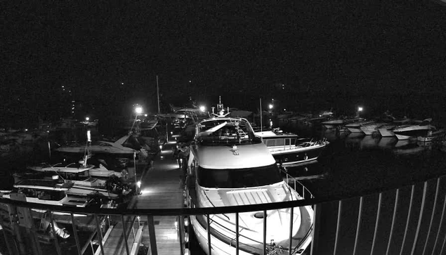 A panoramic view of a marina at night featuring several boats docked in the water. The scene is illuminated by various lights reflecting off the water. In the foreground, a large white boat is prominent, with smaller boats tied alongside. The surrounding area is dimly lit, creating a tranquil nighttime atmosphere. The dock stretching out toward the boats is visible with railings lining the sides.