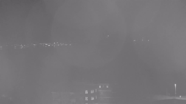 A nighttime webcam view showing a darkened landscape with faint outlines of buildings in the foreground. In the distance, soft lights can be seen, possibly from a city or harbor, reflecting off the water. The overall atmosphere is dim, with a grayish tint and some blurriness, suggesting mist or low visibility conditions.