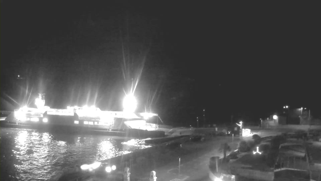 A nighttime scene featuring a brightly lit ferry docked at a waterfront. The ferry is illuminated with strong white lights, reflecting on the dark water. Surrounding the ferry is a dimly lit area with parked vehicles and a few distant lights along the shore. The overall atmosphere is quiet and calm, with no visible movement in the surroundings.