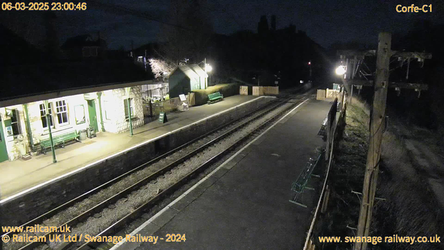 A dimly lit railway station platform at night. On the left, there is a stone building with windows and a green bench in front. A sign saying "Way Out" is visible near the entrance. The platform extends to the right where more green benches are lined up. There are railroad tracks running along the platform, and soft lighting from a nearby lamp illuminates the scene. In the background, there are outlines of buildings and trees in the darkness.