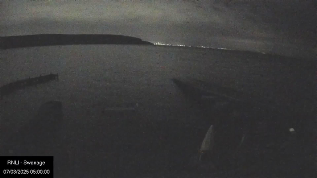 A dark nighttime scene showing a calm body of water with faint ripples. In the background, a distant shoreline is barely illuminated by light, indicating a town or city. A silhouette of a structure or boat is visible in the foreground, with several horizontal lines suggesting the presence of a dock or pier. The sky is overcast, with cloud cover obscuring many stars. The overall atmosphere is quiet and serene.